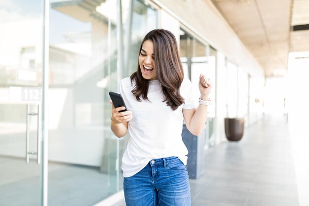 Jeune femme lisant des nouvelles incroyables dans son smartphone et ayant l'air très excitée dans un centre commercial