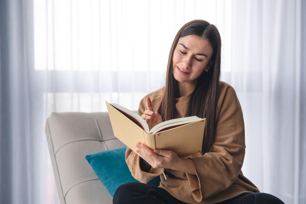 Une jeune femme lit un livre assise sur un fauteuil près de la fenêtre