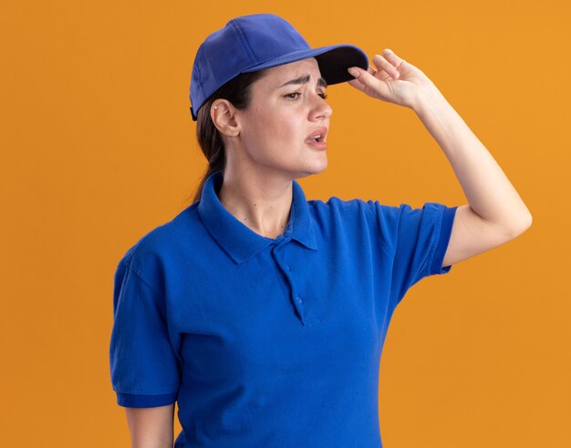 Jeune femme de livraison agacée en uniforme et casquette attrapant la casquette regardant le côté isolé sur le mur orange