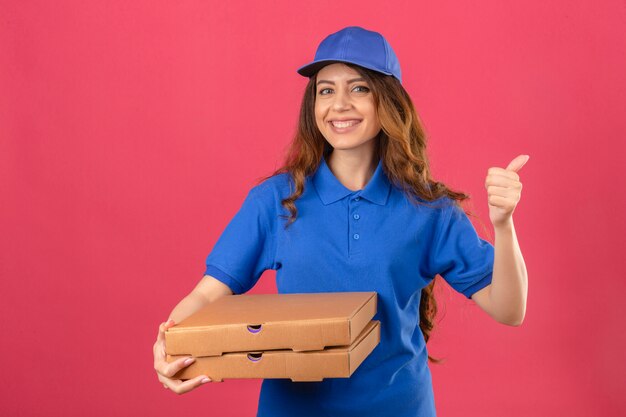 Jeune femme de livraison aux cheveux bouclés portant un polo bleu et une casquette debout avec des boîtes de pizza à la confiance montrant le pouce vers le haut souriant sur fond rose isolé