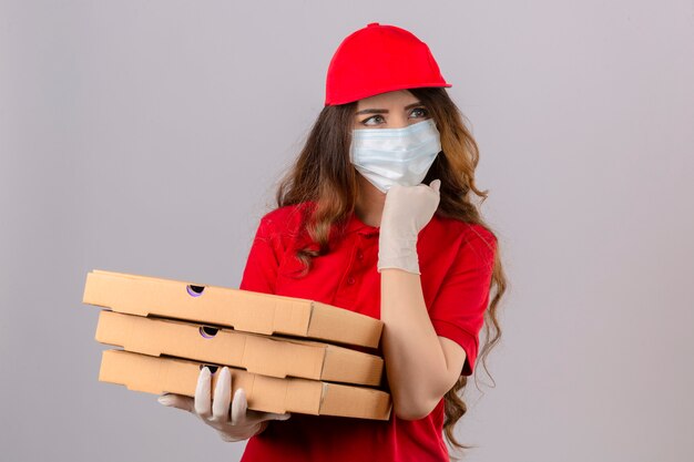 Jeune femme de livraison aux cheveux bouclés portant un polo rouge et une casquette en masque de protection médicale et des gants debout avec des boîtes à pizza pensant concentré sur le doute avec la main sur le menton sur isolé