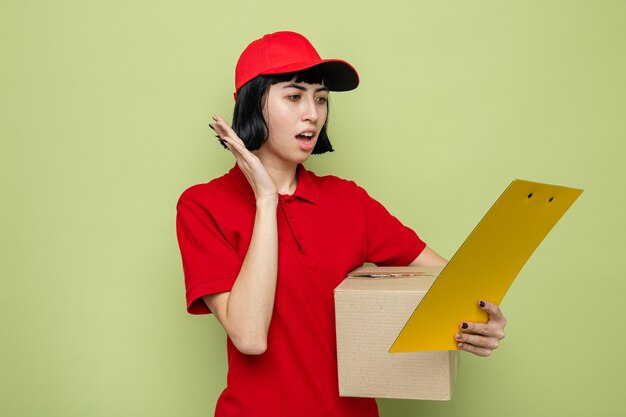 Jeune femme de livraison caucasienne anxieuse tenant une boîte en carton et regardant le presse-papiers