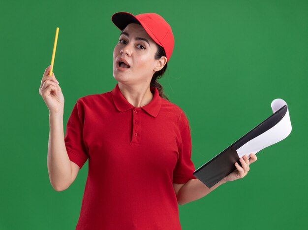 Jeune femme de livraison impressionnée en uniforme et casquette tenant un presse-papiers et un crayon regardant l'avant isolé sur le mur