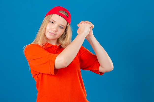 Jeune femme de livraison portant un polo orange et une casquette rouge faisant des gestes avec joint à l'air confiant et fier sur fond bleu isolé