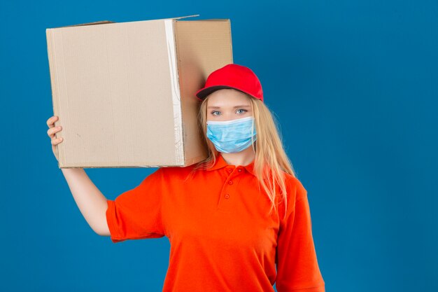 Jeune femme de livraison portant un polo orange et une casquette rouge en masque de protection médicale debout avec une boîte en carton sur l'épaule en regardant la caméra sur fond bleu isolé