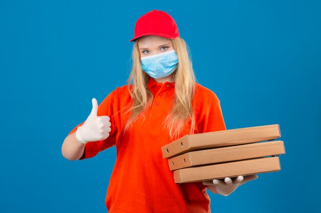 Jeune femme de livraison portant un polo orange et une casquette rouge en masque de protection médicale tenant une pile de boîtes à pizza montrant le pouce vers le haut en souriant sur fond bleu isolé