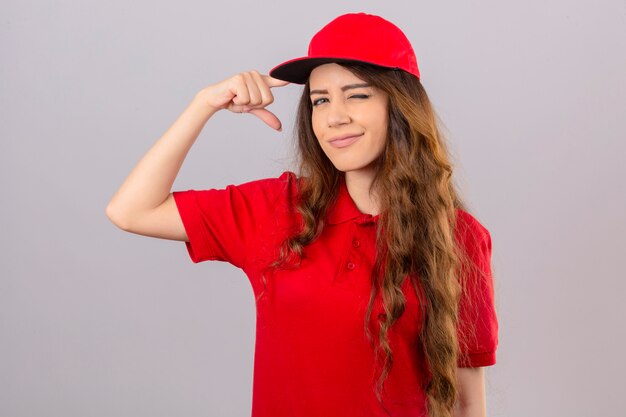 Jeune femme de livraison portant un polo rouge et une casquette avec un clin de œil temple pointant avec le doigt sur fond blanc isolé