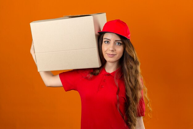 Jeune femme de livraison portant un polo rouge et une casquette debout avec une boîte en carton sur l'épaule à la confiance sur fond orange isolé