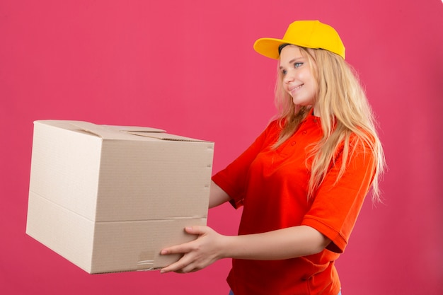 Jeune femme de livraison portant un polo rouge et une casquette jaune donnant une boîte en carton à un client avec sourire sur le visage sur fond rose isolé