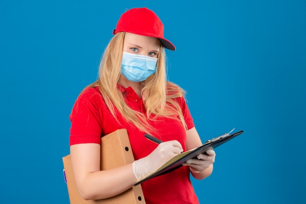 Jeune femme de livraison portant un polo rouge et une casquette en masque de protection médicale debout avec boîte à pizza et presse-papiers écrit à la confiance sur fond bleu isolé