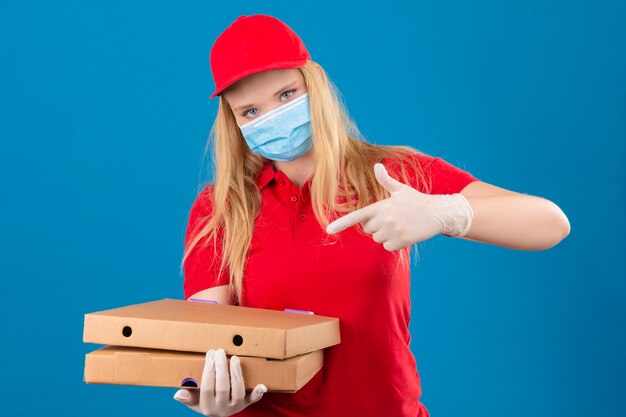 Jeune femme de livraison portant un polo rouge et une casquette en masque de protection médicale debout avec des boîtes à pizza pointant du doigt pour eux regardant la caméra avec un visage sérieux sur dos bleu isolé