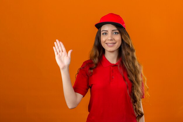 Jeune femme de livraison portant un polo rouge et une casquette souriant joyeusement et joyeusement en agitant la main pour vous accueillir et vous saluer ou dire au revoir sur fond orange isolé