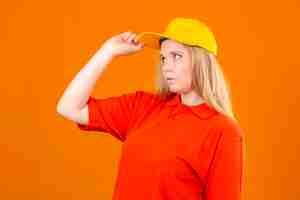 Photo gratuite jeune femme de livraison réfléchie portant un polo rouge et une casquette jaune debout sur le côté pensant avoir des doutes sur fond orange isolé