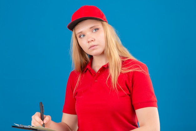 Jeune femme de livraison réfléchie portant un polo rouge et une casquette tenant un presse-papiers avec un stylo à côté sur fond bleu isolé