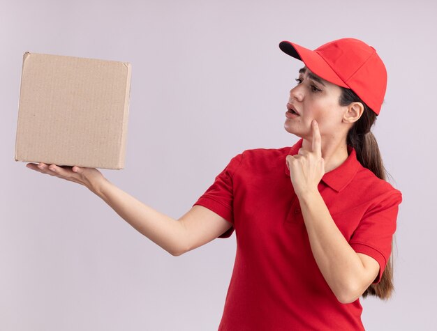 Jeune femme de livraison réfléchie en uniforme et casquette tenant et regardant la boîte à cartes touchant le visage avec le doigt