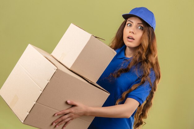 Jeune femme de livraison tendue aux cheveux bouclés portant un polo bleu et une casquette avec des boîtes à la surprise sur fond vert isolé