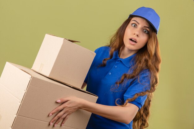 Jeune femme de livraison tendue aux cheveux bouclés portant un polo bleu et une casquette avec des boîtes à la surprise sur fond vert isolé