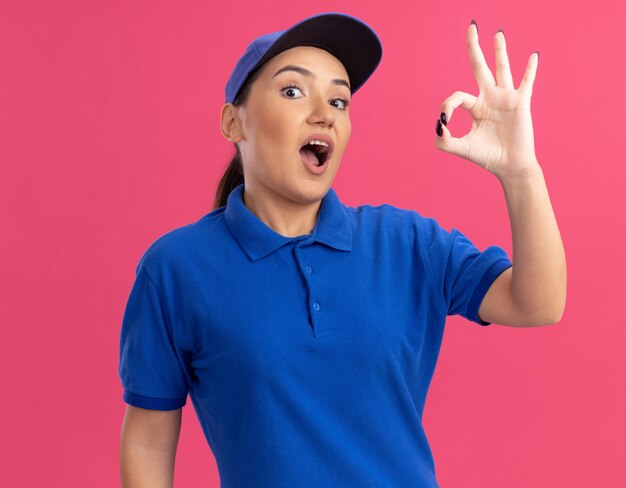 Jeune femme de livraison en uniforme bleu et cap à l'avant heureux et excité montrant signe ok debout sur le mur rose