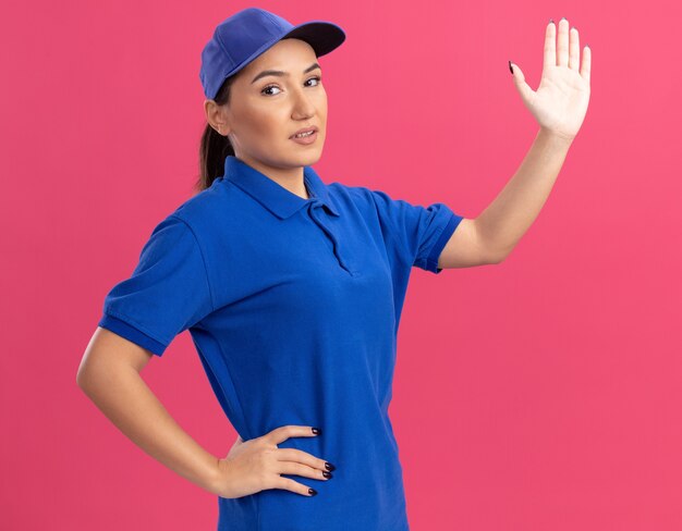 Jeune femme de livraison en uniforme bleu et cap à l'avant souriant confiant levant la main ouverte debout sur le mur rose