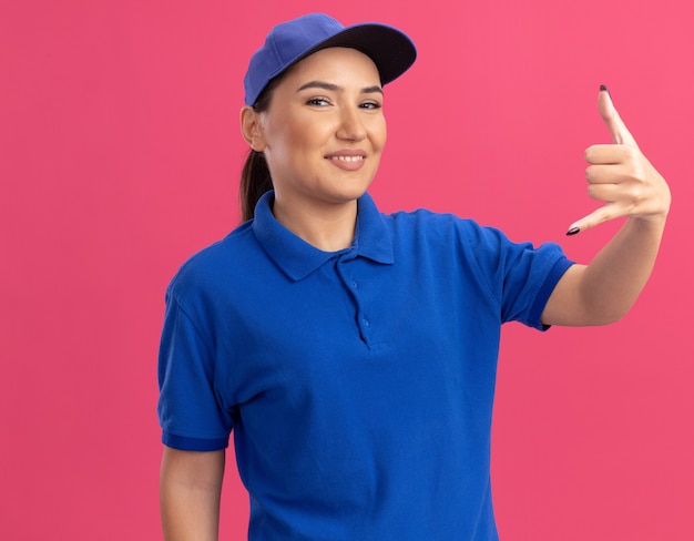 Jeune femme de livraison en uniforme bleu et cap à l'avant souriant joyeusement faisant appelez-moi geste debout sur le mur rose