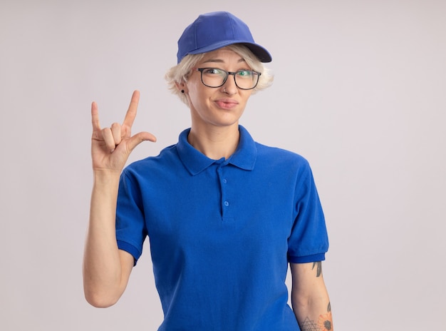 Jeune femme de livraison en uniforme bleu et cap à confus faisant symbole rock debout sur un mur blanc