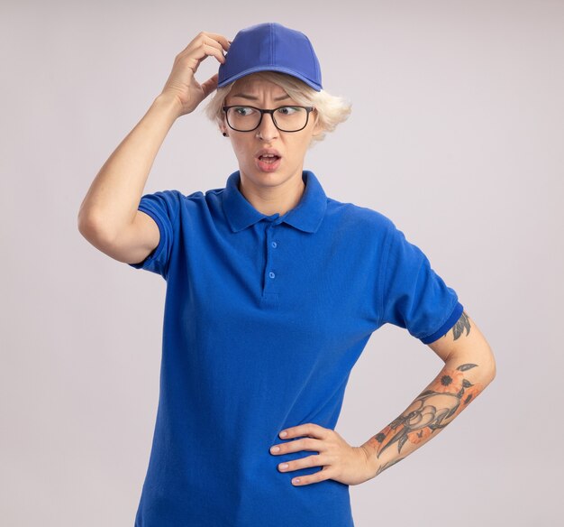 Jeune femme de livraison en uniforme bleu et cap à côté d'être confondu avec la main sur sa tête pour erreur debout sur un mur blanc