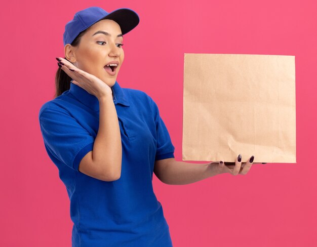 Jeune femme de livraison en uniforme bleu et cap holding paper package regardant étonné et surpris debout sur un mur rose