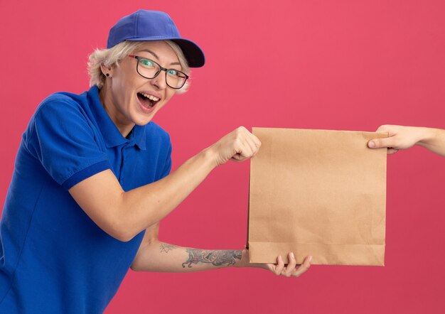 Jeune femme de livraison en uniforme bleu et cap se sentant excité tout en recevant un paquet de papier debout sur un mur rose