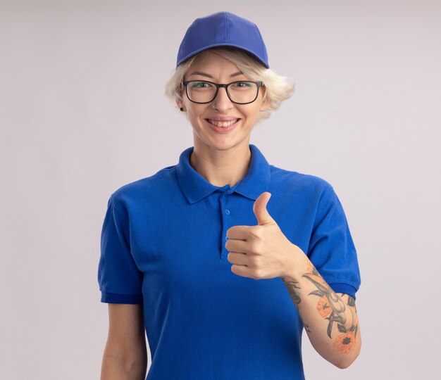 Jeune femme de livraison en uniforme bleu et cap à sourire joyeusement montrant les pouces vers le haut debout sur un mur blanc