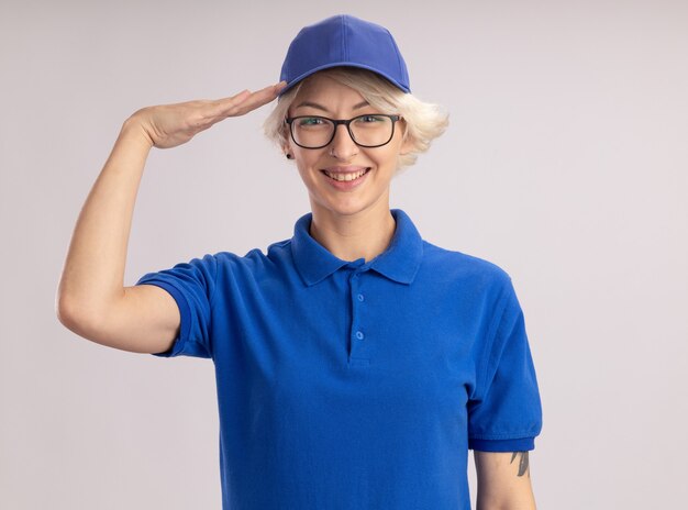 Jeune femme de livraison en uniforme bleu et cap à sourire joyeusement saluant debout sur un mur blanc