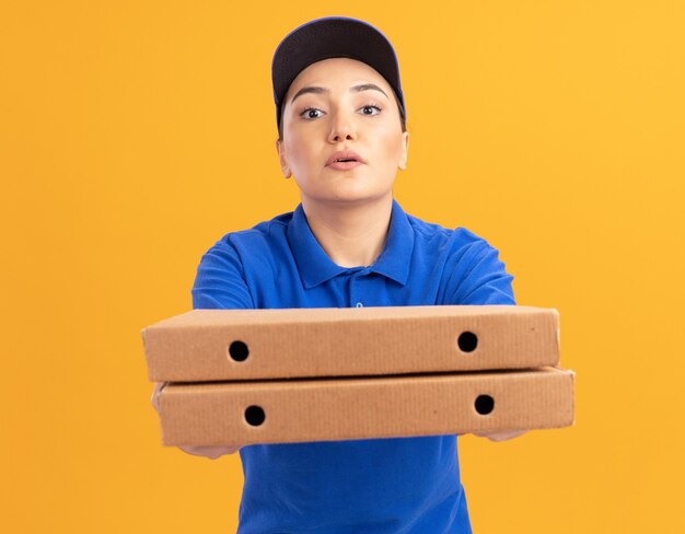 Jeune femme de livraison en uniforme bleu et cap tenant des boîtes de pizza à l'avant avec une expression confiante debout sur un mur orange
