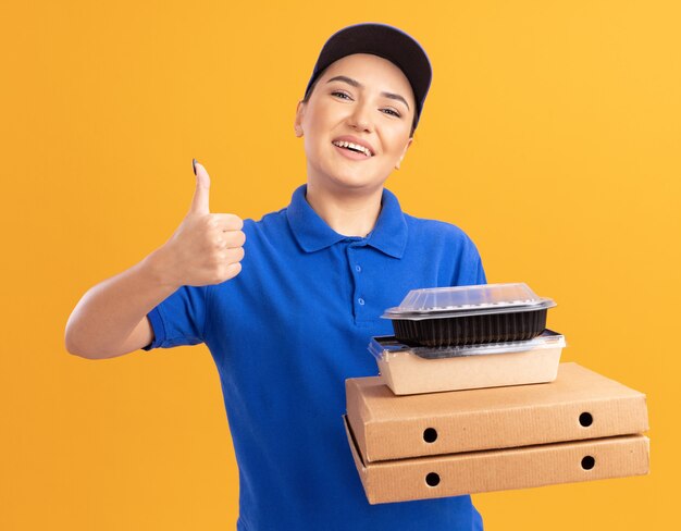 Jeune femme de livraison en uniforme bleu et cap tenant des boîtes de pizza et des emballages alimentaires à l'avant souriant joyeusement montrant les pouces vers le haut debout sur le mur orange