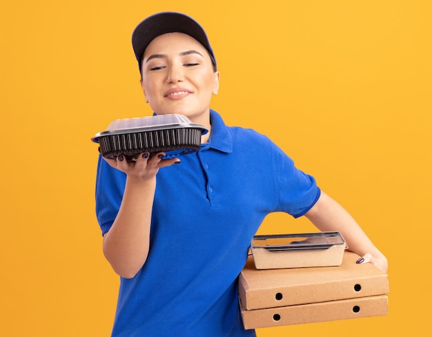 Jeune femme de livraison en uniforme bleu et cap tenant des boîtes de pizza et des emballages alimentaires inhalant un arôme agréable debout sur un mur orange