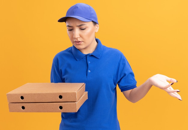 Jeune femme de livraison en uniforme bleu et cap tenant des boîtes de pizza en les regardant être confus debout sur un mur orange