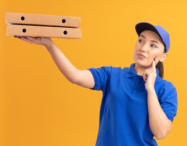 Jeune femme de livraison en uniforme bleu et cap tenant des boîtes de pizza en les regardant intrigués debout sur un mur orange