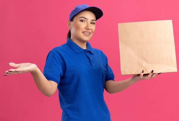 Jeune femme de livraison en uniforme bleu et cap tenant le paquet de papier à l'avant souriant avec visage heureux présentant avec le bras de sa main debout sur le mur rose