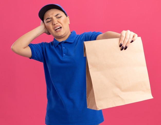 Jeune femme de livraison en uniforme bleu et cap tenant le paquet de papier à la regarder avec une expression confuse d'être mécontent debout sur un mur rose