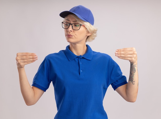 Photo gratuite jeune femme de livraison en uniforme bleu et casquette portant des lunettes à la confiance gesticulant avec les mains, le concept de langage corporel debout sur un mur blanc