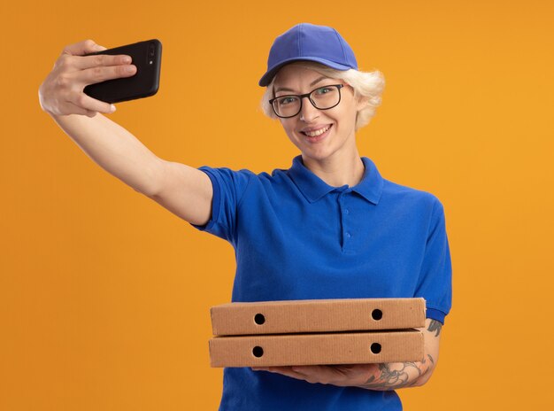 Jeune femme de livraison en uniforme bleu et casquette portant des lunettes tenant des boîtes de pizza à l'aide de smartphone faisant selfie smiling over orange wall