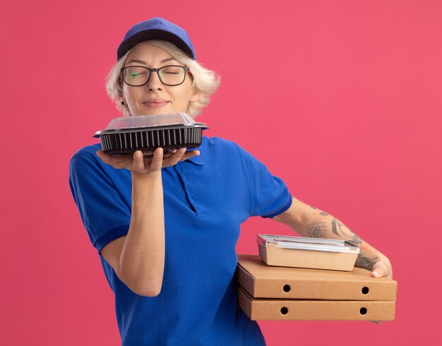 Jeune femme de livraison en uniforme bleu et casquette portant des lunettes tenant des boîtes de pizza et des emballages alimentaires en inhalant un arôme agréable sur un mur rose