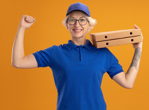 Jeune femme de livraison en uniforme bleu et casquette portant des lunettes tenant des boîtes de pizza levant le poing heureux et gai sur mur orange