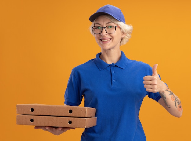 Jeune femme de livraison en uniforme bleu et casquette portant des lunettes tenant des boîtes de pizza montrant les pouces vers le haut souriant sur mur orange