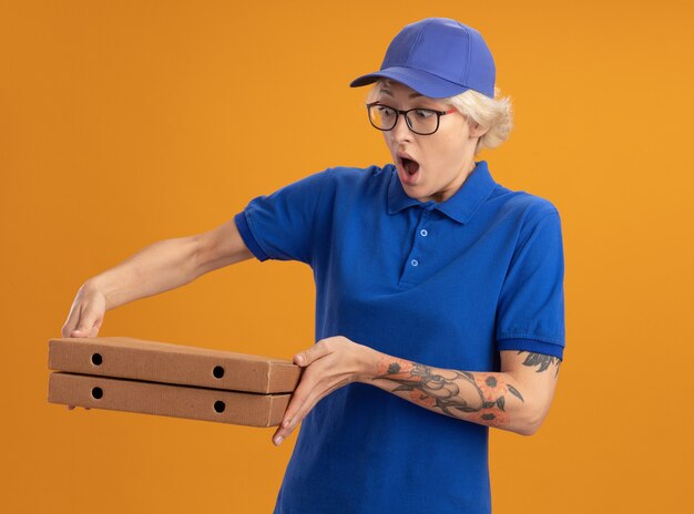 Jeune femme de livraison en uniforme bleu et casquette portant des lunettes tenant des boîtes de pizza en les regardant étonné et surpris sur mur orange