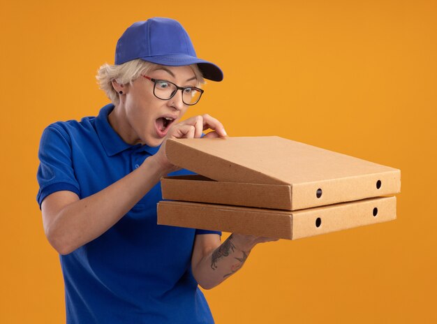 Jeune femme de livraison en uniforme bleu et casquette portant des lunettes tenant des boîtes de pizza en les regardant intriguées sur mur orange