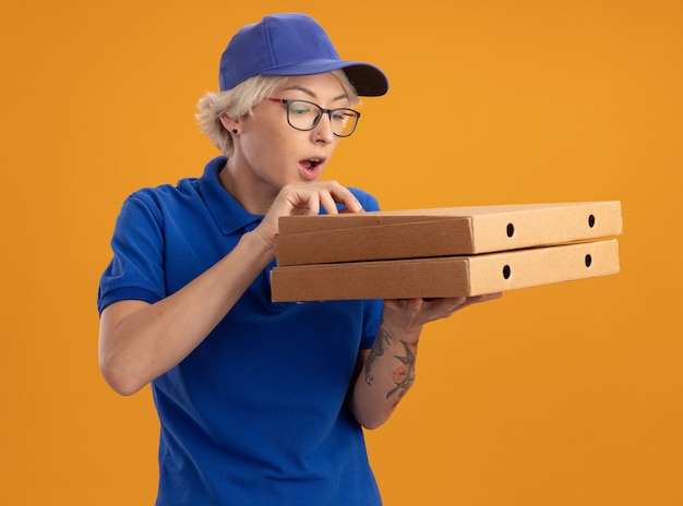 Jeune femme de livraison en uniforme bleu et casquette portant des lunettes tenant des boîtes de pizza en les regardant intriguées sur mur orange
