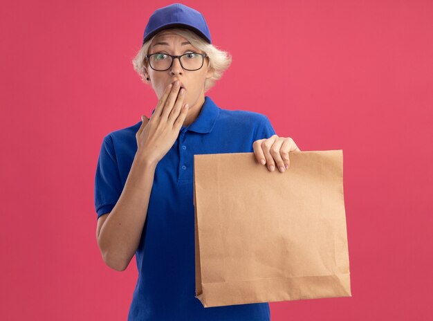 Jeune femme de livraison en uniforme bleu et casquette portant des lunettes tenant un paquet de papier étant choqué couvrant la bouche avec la main sur le mur rose