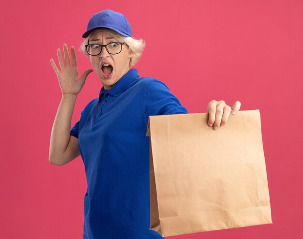 Jeune femme de livraison en uniforme bleu et casquette portant des lunettes tenant un paquet de papier en le regardant dans la panique en levant la main sur le mur rose