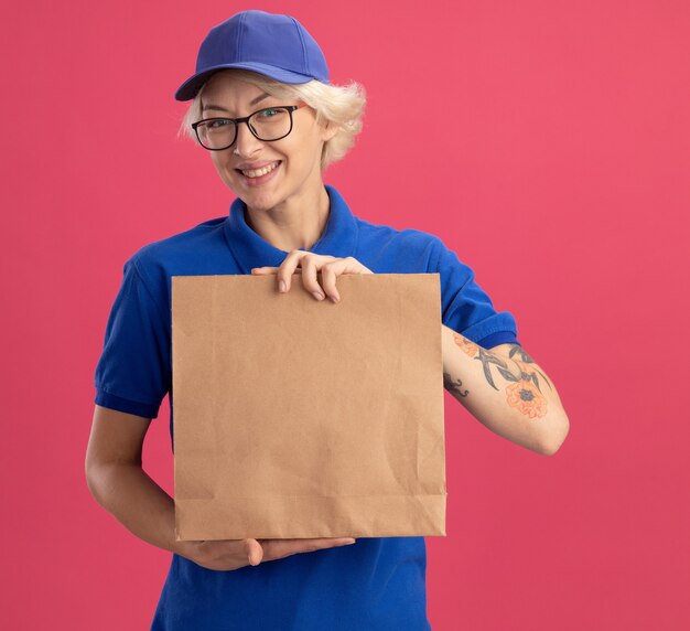 Jeune femme de livraison en uniforme bleu et casquette portant des lunettes tenant un paquet de papier souriant joyeusement sur mur rose