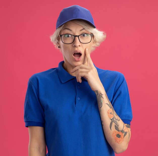 Jeune femme de livraison en uniforme bleu et casquette surpris et étonné avec la bouche grande ouverte sur le mur rose