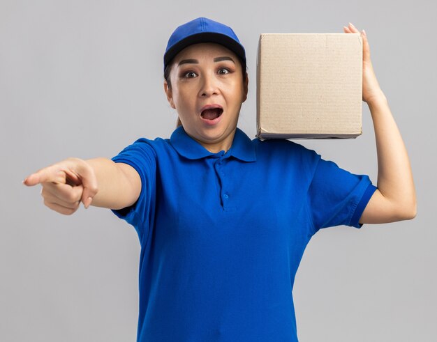 Jeune femme de livraison en uniforme bleu et casquette tenant une boîte en carton à la surprise de pointer avec l'index à la caméra